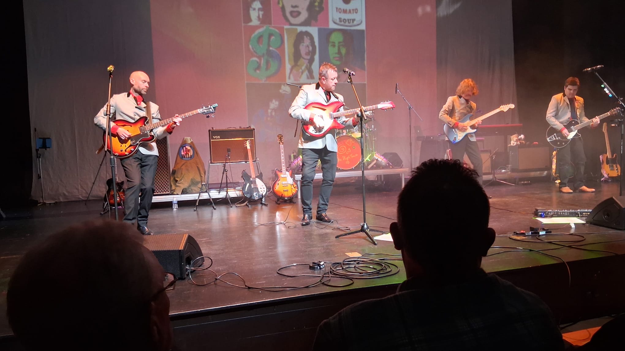 The full five-piece band on stage in matching grey suits, mid-song in front of a Warhol-style pop-art backdrop