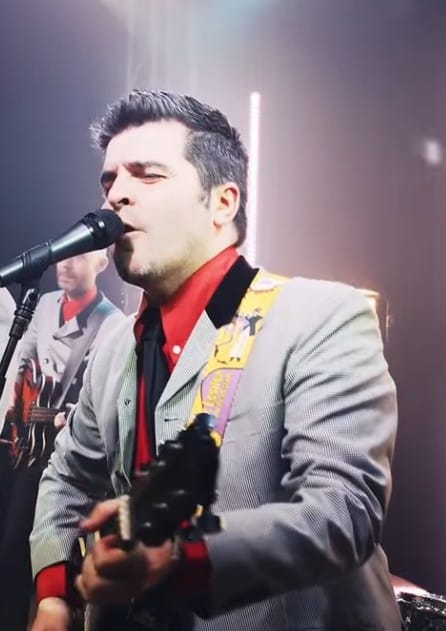 Steve singing into the mic while playing guitar under bright stage lights